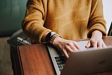 Frau mit gelbem Pullover sitzt an Holztisch vor aufgeklapptem Laptop.