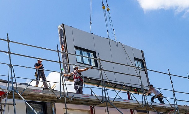 Energiesprong: serielle Sanierung in Longueau/Frankreich (Baugerüst mit Bauarbeitern und Fassadenelement an Kran-Ketten).